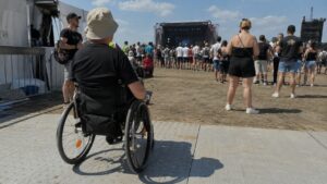 Personne en fauteuil roulant assistant à un festival de musique en plein air lors d’une journée ensoleillée, devant une scène de concert avec une foule de festivaliers, illustrant l’accessibilité des événements culturels aux personnes à mobilité réduite.
