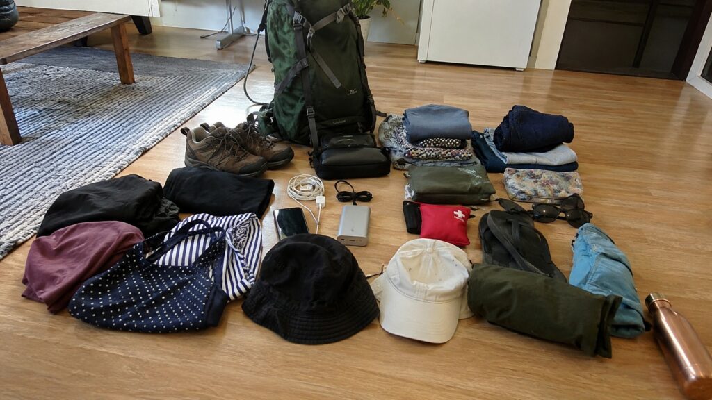 Photo Panasonic Lumix S5 montrant une checklist complète pour le festival L'Art de Rien avec grand sac à dos de randonnée vert, vêtements pliés, chaussures de marche, batterie externe, gourde, lunettes de soleil, casquettes, trousse de secours et accessoires de voyage disposés sur un parquet en intérieur avant le départ.